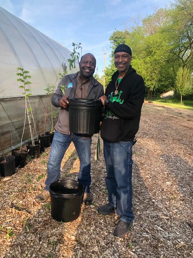 Two smiling men holding a black plant pot between them on a wood-chip path beside a greenhouse, with young trees in pots behind them